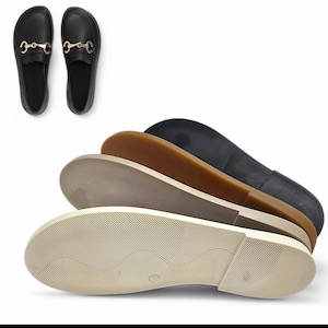 May include: Black loafers with gold-colored horsebit detail, displayed with the soles of several pairs of shoes in various colors: beige, brown, and black. The soles have a textured, ridged pattern.
