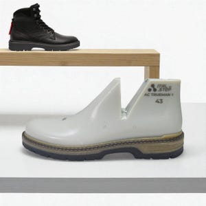 Puede incluir: Una bota de cuero negro con suela gruesa se encuentra en una estantería de madera sobre una horma blanca. La horma tiene el texto "ITAL STEP AC TRUEMAN Y 43" impreso. La bota tiene una lengüeta roja.