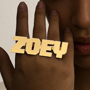 Pode incluir: Um anel dourado com o nome "ZOEY" em letras maiúsculas. O anel é usado em um dedo, com a mão e parte do rosto visíveis. O anel é o ponto focal, com um fundo simples e neutro. O anel é brilhante.