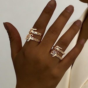 May include: Two gold rings, each bearing a name in cursive script, are shown on a hand. The names are "Funm" and "Damin". The rings are set against a plain background, with a white blazer in view.