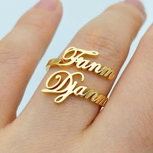 May include: A gold-coloured, double-band ring with the names "Fann" and "Diann" in a cursive font. The ring is worn on a finger, showcasing the personalised jewellery design. The ring is made of a shiny, metallic material.