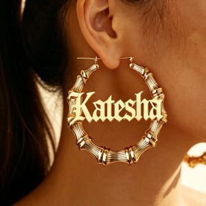 May include: A large gold-toned hoop earring featuring a bamboo-inspired texture and a central horizontal bar displaying the name Katesha in a stylized gothic font. The circular earring is polished with a reflective metallic finish.