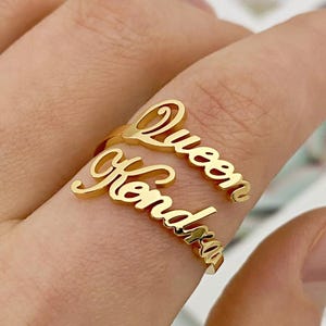 Puede incluir: Anillo de oro con los nombres "Queen" y "Kendra" entrelazados.