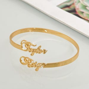 Op de afbeelding: Een goudkleurige, open manchetarmband met de namen "Taylor" en "Reign" in een cursief lettertype. De armband is gemaakt van glanzend metaal en wordt op een wit oppervlak weergegeven.