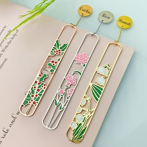 Puede incluir: Tres marcapáginas decorativos de metal con diseños florales. Uno es dorado con acebo y bayas, otro es plateado con claveles rosas y el tercero es dorado con lirios de los valles. Cada uno tiene una cadena y una etiqueta personalizada con un nombre.