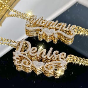 Könnte beinhalten: Zwei goldfarbene Namensketten, eine mit der Aufschrift "Denise" und die andere mit "Monique". Jede Halskette hat ein kursives Namensdesign mit Herzakzent, das mit kleinen, funkelnden Steinen besetzt ist. Die Halsketten werden auf einer reflektierenden Oberfläche präsentiert.