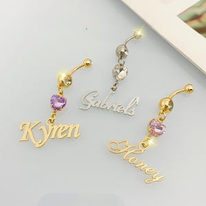 May include: Three gold belly button rings with a dangling charm. The charms are a heart with a purple rhinestone, a heart with a pink rhinestone, and a cursive name "Honey". The other two rings have the names "Kyren" and "Gabriels" dangling from them.