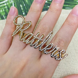 Può includere: Un anello dorato per più dita che riporta il nome Kathleen in corsivo. La superficie delle lettere è incastonata con numerosi piccoli strass chiari e scintillanti che catturano la luce, creando un aspetto luminoso e molto riflettente.