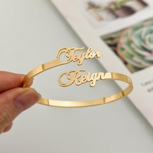 May include: Gold-toned bangle bracelet with two names, "Taylor" and "Reign", written in a cursive script.