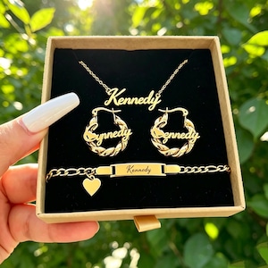 May include: A gold-toned jewelry set featuring a necklace, two hoop earrings, and a bracelet, all personalized with the name Kennedy in script. The bracelet includes a small heart charm and a rectangular nameplate, presented in a square box.