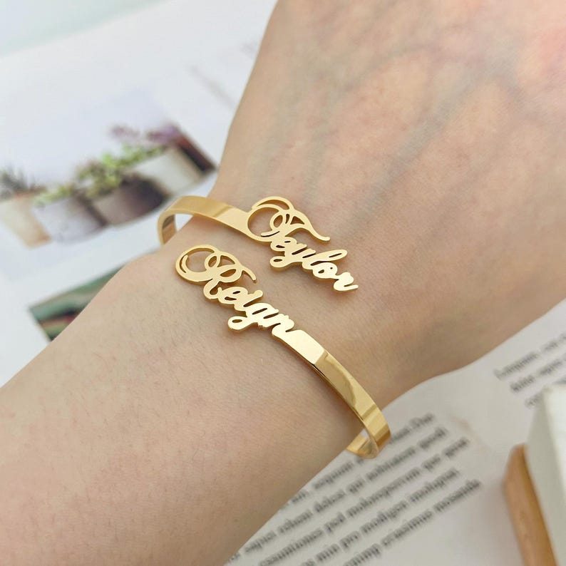 May include: Gold bangle bracelet with two personalized name charms. The names "Taylor" and "Regan" are spelled out in cursive.