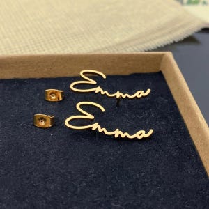 May include: A pair of gold-toned stud earrings featuring the name Emma written in a cursive script font. The set includes two individual earring pieces and two matching gold-toned earring backs, all displayed on a dark blue fabric surface.