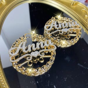 Puede incluir: Pendientes de aro dorados con el nombre "Anna" escrito en una fuente brillante similar a diamantes. El nombre está centrado sobre un diseño en forma de corazón. Los aros tienen un diseño de cuerda trenzada.