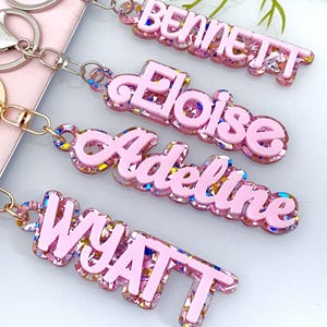 Puede incluir: Cuatro llaveros personalizados con los nombres: Bennett, Eloise, Adeline y Wyatt. Cada llavero presenta un diseño rosa claro en capas con detalles de purpurina y un llavero dorado.