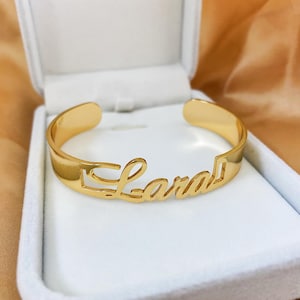 May include: A gold cuff bracelet, inscribed with the name "Lara" in a cursive font. The bracelet is presented in a white jewellery box with a velvet lining. The background is a soft, golden fabric.