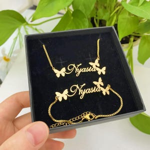 May include: A gold-coloured jewellery set comprising a necklace and bracelet, both inscribed with the name "Nyasia" in cursive script. Butterfly designs are integrated into the pieces. The set is displayed in a black presentation box.
