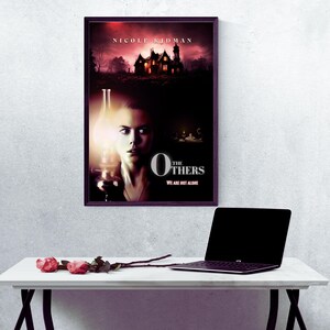 May include: Movie poster for the film "The Others" starring Nicole Kidman. The poster features a woman holding a lantern in front of a dark, ominous house. The text "The Others" is in large, white letters with the tagline "We are not alone" below.