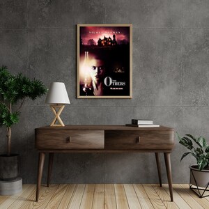 May include: A movie poster for the film "The Others" starring Nicole Kidman. The poster features a woman with a pale complexion and dark hair standing in front of a large, dark house. The text "The Others" is written in white letters on a black background.
