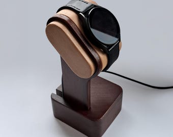 Puede incluir: Un reloj inteligente negro con una correa de cuero descansa sobre un soporte de reloj de madera. El soporte es de madera marrón oscuro y tiene un diseño curvo para acunar el reloj. Un cable de carga negro está conectado al soporte.