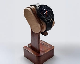 Puede incluir: Un reloj inteligente negro con una correa de cuero negro descansa sobre un soporte de reloj de madera. El soporte es de madera marrón oscuro con una cuna marrón claro. La esfera del reloj muestra la hora.