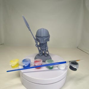 May include: A gray Mandalorian figurine holding a spear, standing on a white platform. Paint pots in yellow, blue, red, green, white, and black are arranged in front of the figure, along with a blue-handled paintbrush. The figurine is a DIY craft project.