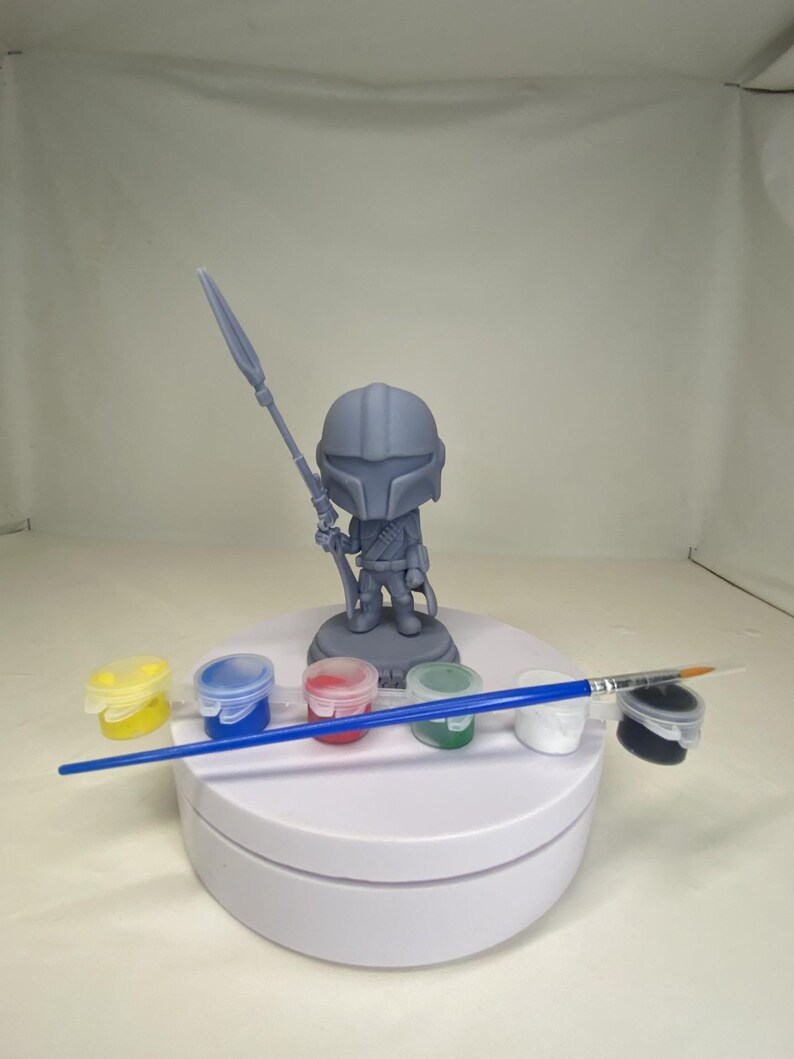 May include: A gray Mandalorian figurine holding a spear, standing on a white platform. Paint pots in yellow, blue, red, green, white, and black are arranged in front of the figure, along with a blue-handled paintbrush. The figurine is a DIY craft project.