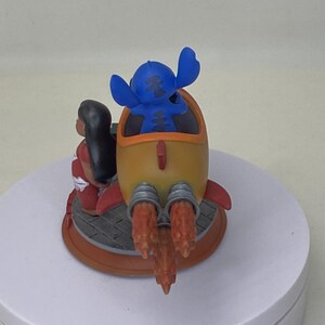 May include: A figurine featuring Stitch in a yellow rocket with red accents, and Lilo in a red dress. The rocket has orange flames and is on a circular base. The characters are from the animated movie Lilo & Stitch.