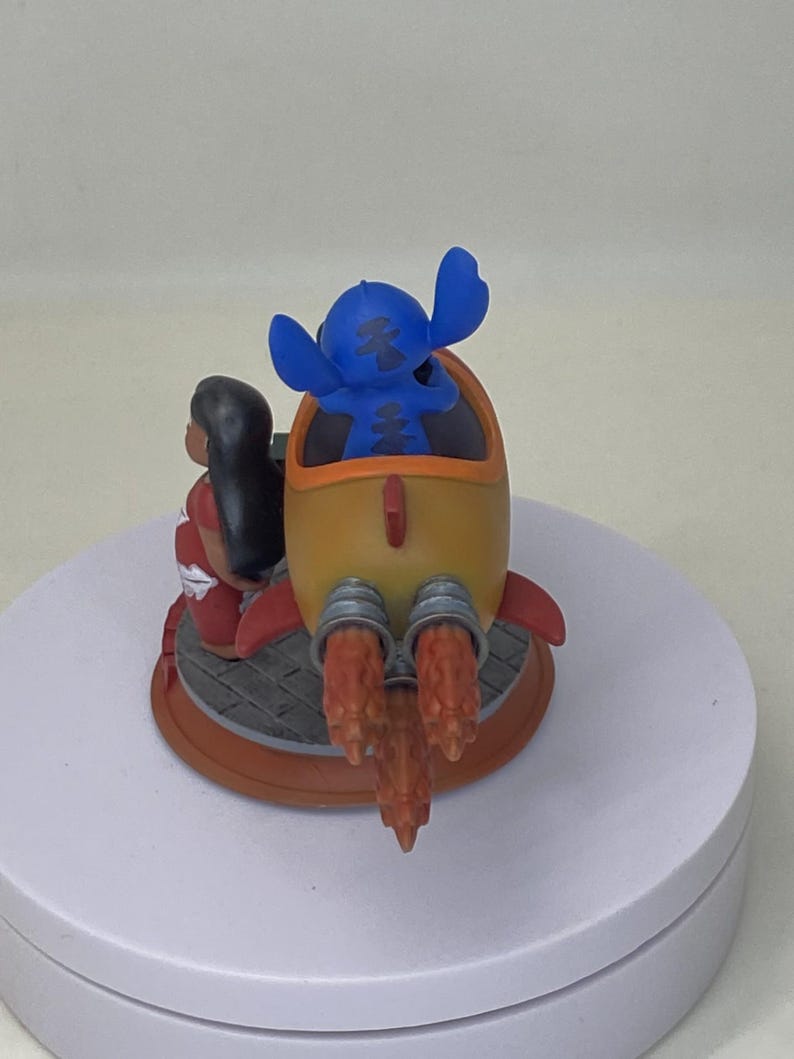 May include: A figurine featuring Stitch in a yellow rocket with red accents, and Lilo in a red dress. The rocket has orange flames and is on a circular base. The characters are from the animated movie Lilo & Stitch.