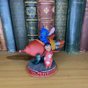 May include: A figurine featuring Stitch in a red rocket ship and Lilo in a red dress. The base reads "Lilo & Stitch". The figurine is set against a backdrop of antique books with various titles and colors.