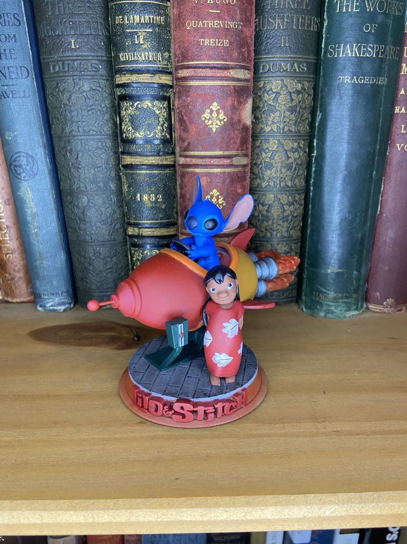 May include: A figurine featuring Stitch in a red rocket ship and Lilo in a red dress. The base reads "Lilo & Stitch". The figurine is set against a backdrop of antique books with various titles and colors.