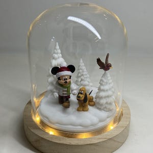 May include: A decorative snow globe featuring Mickey Mouse, Pluto, and a bird in a winter scene. The figures are surrounded by white trees and snow, under a glass dome with a wooden base. The base has small lights.
