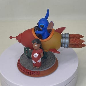 May include: A figurine featuring Stitch in a rocket ship with Lilo. The rocket is red and yellow with orange flames. Stitch is blue and Lilo is wearing a red dress. The base reads "STITCH" in red.