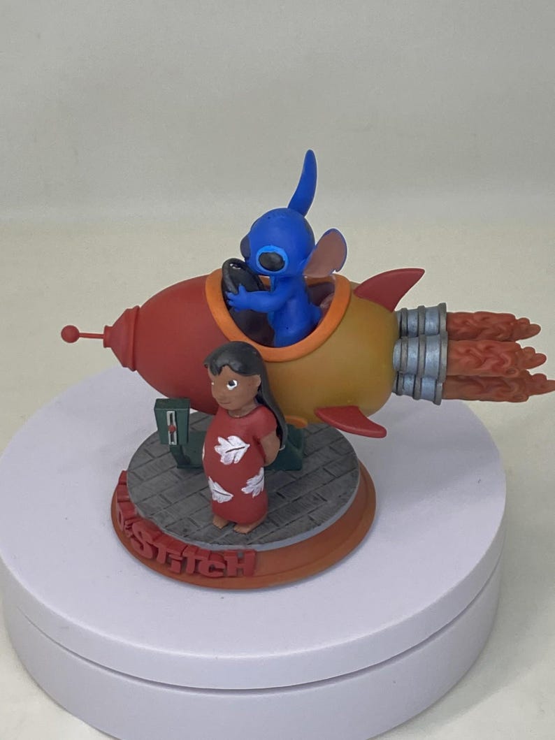 May include: A figurine featuring Stitch in a rocket ship with Lilo. The rocket is red and yellow with orange flames. Stitch is blue and Lilo is wearing a red dress. The base reads "STITCH" in red.
