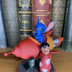 May include: A figurine featuring the character Stitch from Lilo & Stitch in a red and yellow rocket ship, with Lilo standing nearby. The base is a brick pattern with a red border. The background includes antique books.