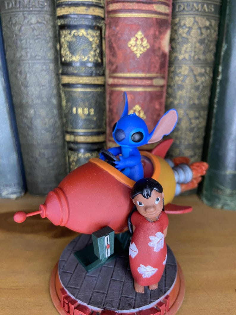 May include: A figurine featuring the character Stitch from Lilo & Stitch in a red and yellow rocket ship, with Lilo standing nearby. The base is a brick pattern with a red border. The background includes antique books.
