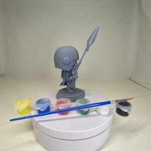 May include: A gray figurine of a character holding a spear, with a helmet and armor. The figurine is on a white rotating stand, surrounded by small paint pots in yellow, blue, red, green, white, and black, and a blue-handled paintbrush.