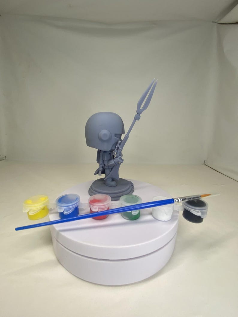 May include: A gray figurine of a character holding a spear, with a helmet and armor. The figurine is on a white rotating stand, surrounded by small paint pots in yellow, blue, red, green, white, and black, and a blue-handled paintbrush.
