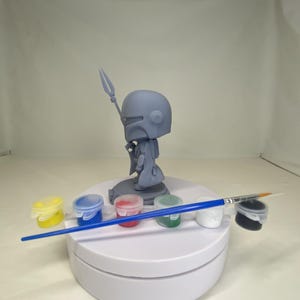 May include: A gray Mandalorian figurine with a spear, displayed on a white rotating stand. The stand also holds small paint pots in yellow, blue, red, green, white, and black, along with a blue-handled paintbrush.