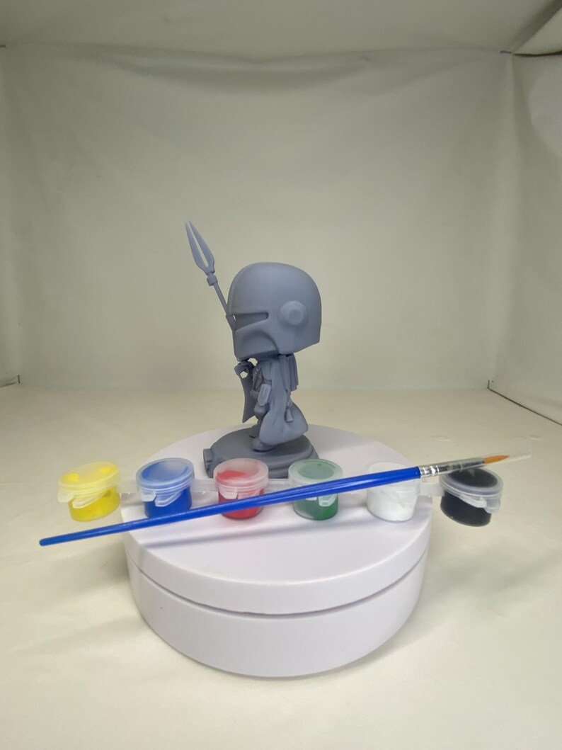 May include: A gray Mandalorian figurine with a spear, displayed on a white rotating stand. The stand also holds small paint pots in yellow, blue, red, green, white, and black, along with a blue-handled paintbrush.
