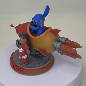 May include: A figurine featuring Stitch in a rocket ship with Lilo standing nearby. The rocket is red and yellow with orange flames. Lilo wears a red dress. The base is brown with the word "Stitch".