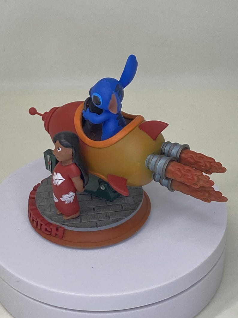 May include: A figurine featuring Stitch in a rocket ship with Lilo standing nearby. The rocket is red and yellow with orange flames. Lilo wears a red dress. The base is brown with the word "Stitch".