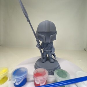 May include: A gray Mandalorian figurine holding a spear, standing on a circular base. The figurine is surrounded by small paint pots in yellow, blue, red, and green, along with a blue paintbrush. This is a DIY craft kit.