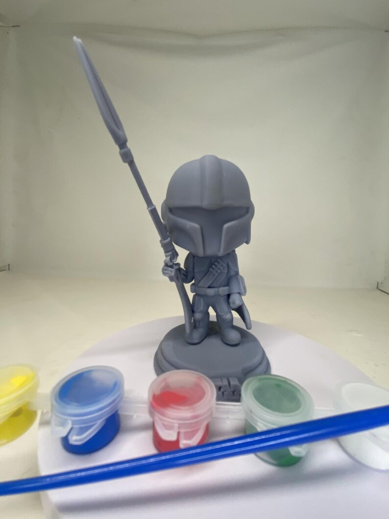May include: A gray Mandalorian figurine holding a spear, standing on a circular base. The figurine is surrounded by small paint pots in yellow, blue, red, and green, along with a blue paintbrush. This is a DIY craft kit.