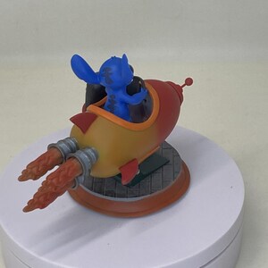 May include: A blue Stitch figurine in a red, yellow, and orange rocket ship. The rocket has silver exhaust ports and a small red antenna. The figurine is on a white rotating display stand.