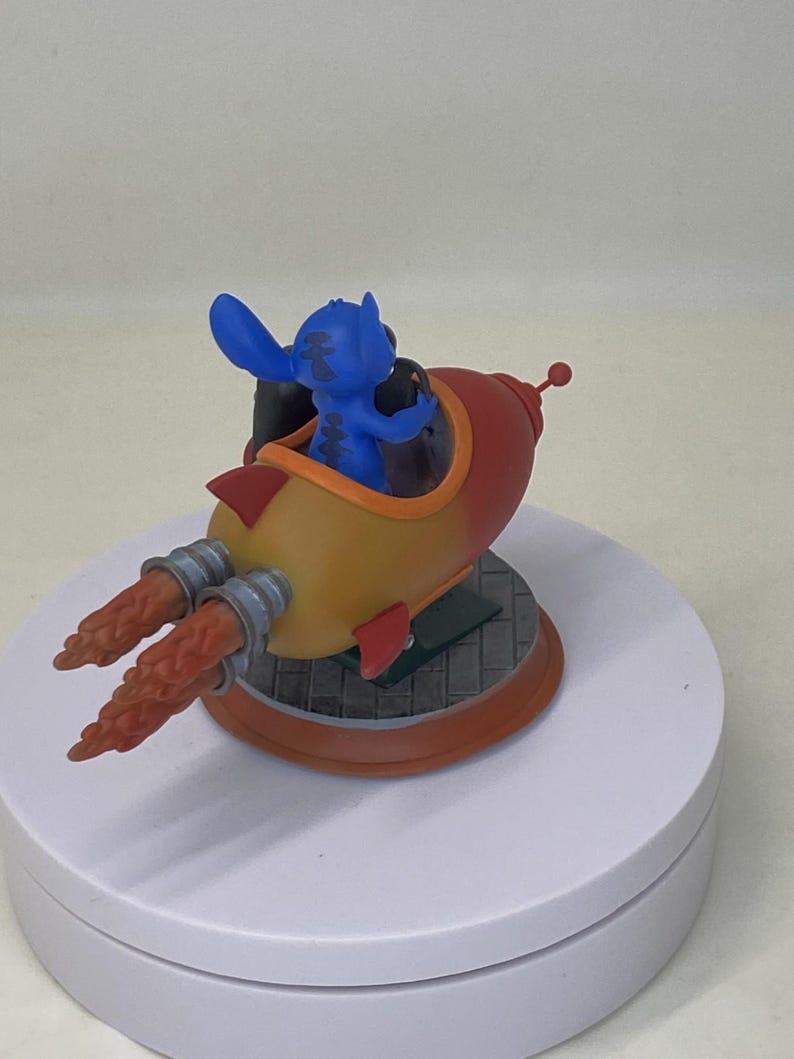 May include: A blue Stitch figurine in a red, yellow, and orange rocket ship. The rocket has silver exhaust ports and a small red antenna. The figurine is on a white rotating display stand.