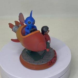 May include: A figurine featuring the character Stitch from Lilo & Stitch in a red and yellow rocket ship. Stitch is driving the ship, with Lilo standing beside him. The base is brown with the word "Lilo" on it.