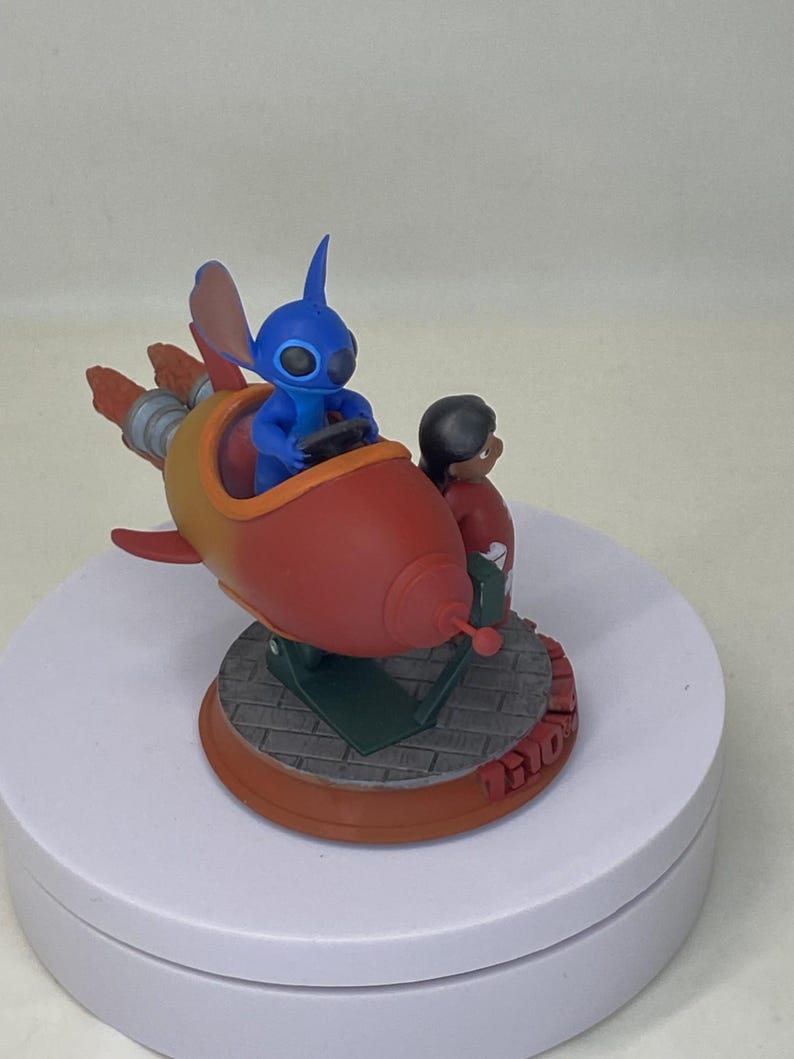 May include: A figurine featuring the character Stitch from Lilo & Stitch in a red and yellow rocket ship. Stitch is driving the ship, with Lilo standing beside him. The base is brown with the word "Lilo" on it.