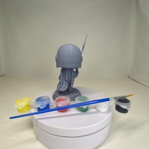 May include: A gray Mandalorian figure with a spear stands on a white platform. Six small paint pots in yellow, blue, red, green, white, and black are arranged in front of the figure. A blue paintbrush rests across the pots.