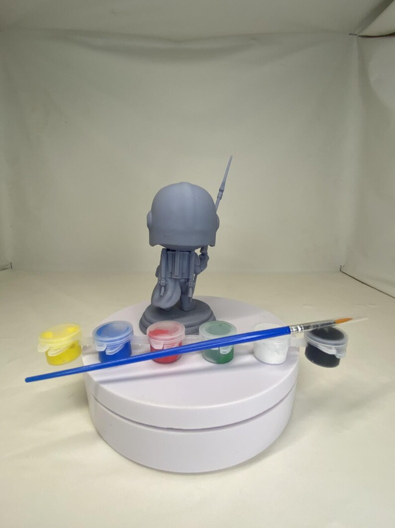 May include: A gray Mandalorian figure with a spear stands on a white platform. Six small paint pots in yellow, blue, red, green, white, and black are arranged in front of the figure. A blue paintbrush rests across the pots.