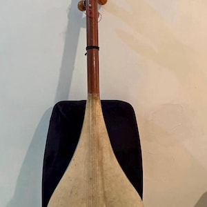 Könnte beinhalten: Ein traditionelles Saiteninstrument mit langem Hals und birnenförmigem Korpus. Das Instrument ist aus Holz gefertigt und hat eine hellfarbige, gespannte Membran auf dem Korpus. Es liegt auf einem schwarzen Stuhl.